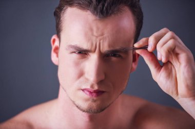 Çıplak gövdeli yakışıklı bir adam gri bir arka planda kaşlarını yoluyor..