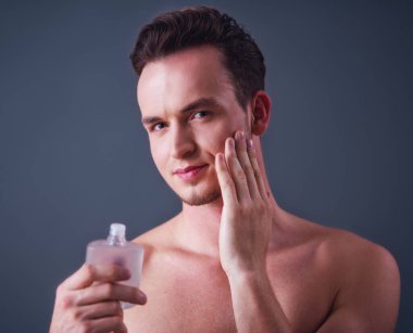 Yakışıklı genç tıraş losyonu holding, kameraya bakıyor ve gülümsüyor, gri arka plan üzerinde