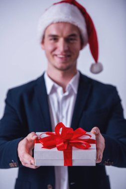 Takım elbise ve Santa şapka yakışıklı adam kameraya bakıyor ve gülümsüyor, üzerinde beyaz izole bir hediye kutusu tutuyor