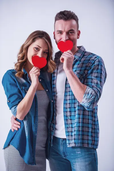 カジュアルな服で美しい若いカップルが つ赤い紙のハート カメラ目線と笑顔 白で隔離を保持しています ストックフォト