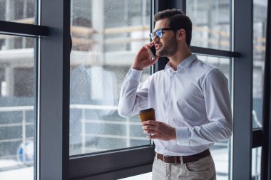 Gözlük yakışıklı işadamı cep telefonda konuşurken ve ofis pencereden dışarı bakarken kahve içme