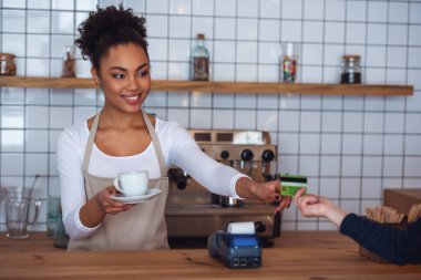 Güzel Afro Amerikan barista önlük bir istemciye, kredi kartı ve sayaç bar, dururken gülümseyerek veriyor