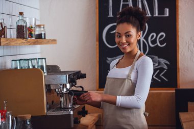 Güzel Afro Amerikan barista önlük kameraya bakarak ve bir kahve makinesi kullanarak kahve yaparken gülümseyen
