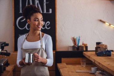Güzel Afro Amerikan barista önlük bir fincan süt Holding'in, uzağa bakarak ve bir kahve makinesi yakın dururken gülümseyen
