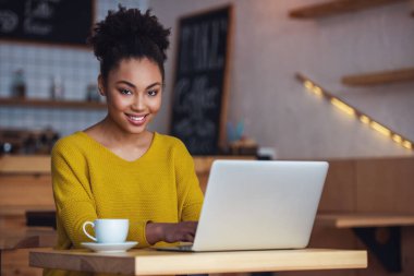 Afro Amerikan Kız güzel rahat kıyafetler bir dizüstü bilgisayar kullanarak, kameraya bakıyor ve kafede otururken gülümseyen