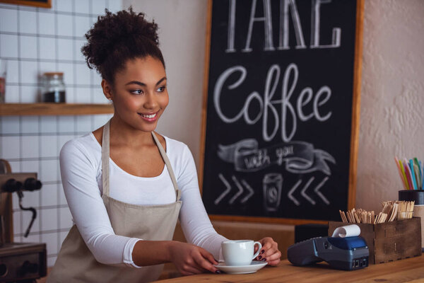 Beautiful Afro American barista in apron is holding a cup and smiling while standing at bar counter