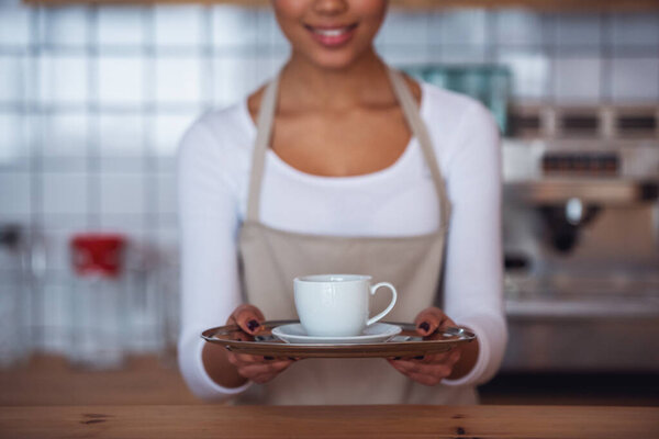 Cropped image of beautiful Afro American barista in apron holding a cup and smiling while standing at bar counter