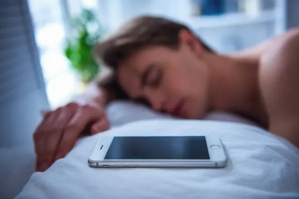 Handsome young man is sleeping in his bed, his smartphone is on the pillow near, phone in focus