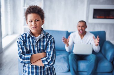 Yakışıklı çocuk kollarını kavuşturmuş ve suç işleyerek kameraya bakarken babası çığlık atıyor, arka planda koltukta oturuyor.