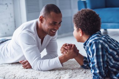 Mutlu baba ve oğul gündelik giysiler içinde bilek güreşi yapıyor ve evde halının üzerinde yatarken gülümsüyorlar.