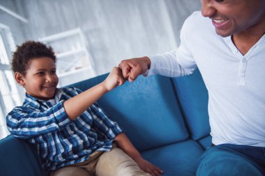 Mutlu baba ve oğul günlük giysiler içinde evde kanepede otururken yumruk tokuşturup gülümsüyorlar.