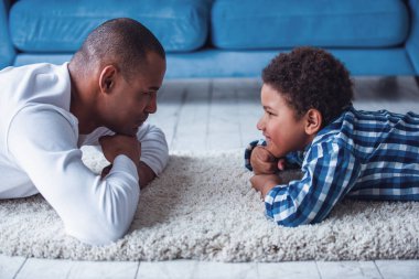 Mutlu baba ve oğul günlük giysiler içinde birbirlerine bakıyorlar ve evde halının üzerinde yatarken gülümsüyorlar.