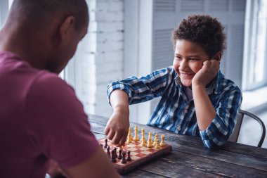 Yakışıklı baba ve oğul gündelik giysiler içinde satranç oynarken evde birlikte vakit geçiriyorlar, çocuk gülümsüyor.