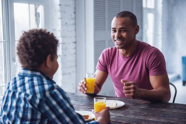 Yakışıklı baba ve oğul gündelik giysiler içinde evde birlikte ahşap masada yiyorlar, adam gülümsüyor.