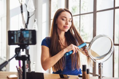 Evde kameraya ateş ederken saç ütüsü kullanan güzel bir kadın blogcu.