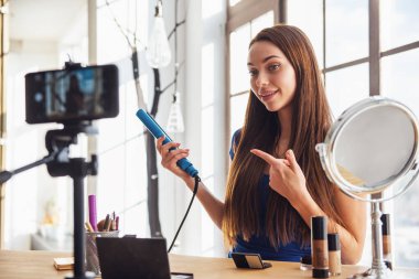 Evde kameraya ateş ederken saç ütüsü kullanan güzel bir kadın blogcu.