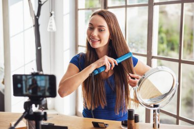 Evde kameraya ateş ederken saç ütüsü kullanan güzel bir kadın blogcu.