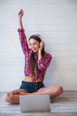 Uzun saçlı genç ve güzel bir kızın portresi dizüstü bilgisayar kullanır ve yerde otururken kulaklıkla müzik dinler