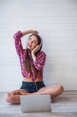 Uzun saçlı genç ve güzel bir kızın portresi dizüstü bilgisayar kullanır ve yerde otururken kulaklıkla müzik dinler