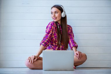 Uzun saçlı genç ve güzel bir kızın portresi dizüstü bilgisayar kullanır ve yerde otururken kulaklıkla müzik dinler