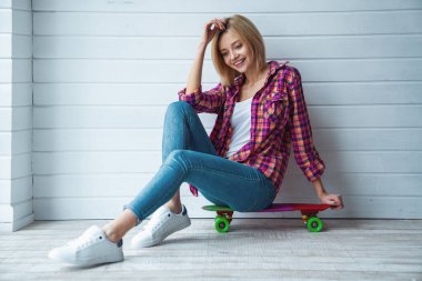 Kaykayın üzerinde oturan ve arka planda kameraya gülümseyen neşeli güzel sarışın kızın portresi.