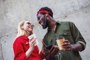 Genç, neşeli bir çift, bir adam ve sarışın bir kadın gri bir duvarın arkasında duruyor, kulaklıkla müzik dinliyor ve kahve içiyorlar.