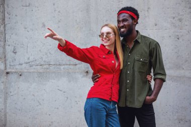 Genç, neşeli bir çift, bir adam ve sarışın bir kadın, gri bir duvarın arka planında sarılıyorlar ve kızların işaret ettiği yere bakıyorlar.