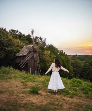 Retro tarzı. Ormanın arka planında beyaz elbiseli, siyah şık saçlı, genç ve güzel bir kız, bir değirmen ve kırmızı bir gün batımı. Sırtı kameraya dönük duruyor..