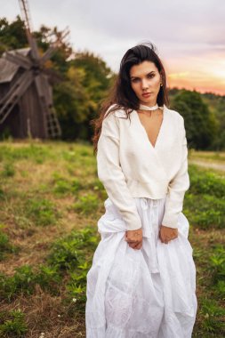 Retro tarzı. Ormanın arka planında beyaz bir elbise, değirmen ve kırmızı gün batımı giymiş siyah şık saçlı güzel bir kızın portresi..