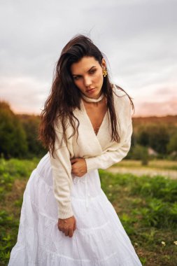 Retro tarzı. Siyah şık saçlı, beyaz elbiseli güzel bir kızın uzun bir portresi bir ormanın arka planında ve kırmızı bir günbatımında..