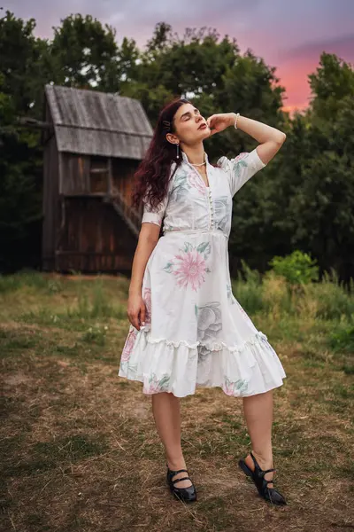 stock image Retro style. Young beautiful girl with black chic hair in a white dress on the background of a forest, a mill and a red sunset.