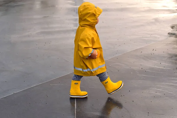 雨の日に黄色いレインコートとゴムブーツを着た小さな子供が水たまりを通って行きます 悪天候の時にアウトドアを楽しむ — ストック写真