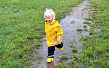 Yağmurlu bir sonbahar gününde çamurlu su birikintisini keşfeden küçük bir çocuk. Yağmurluk giymiş küçük bir çocuk ve açık havada sarı yağmurluk. Çocuklar için sezonluk açık hava aktiviteleri