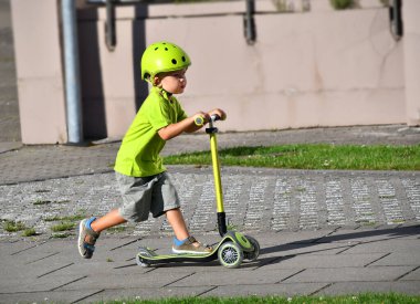 3-4 yaşlarında bir çocuk scooter kullanıyor. Güneşli yaz gününde banliyö sokağında oynayan bir çocuk. Çocuklar için güvenli kask. Sağlıklı açık hava aktivitesi.