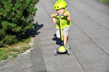 3-4 yaşlarında bir çocuk scooter kullanıyor. Güneşli yaz gününde banliyö sokağında oynayan bir çocuk. Çocuklar için güvenli kask. Sağlıklı açık hava aktivitesi.