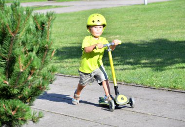 3-4 yaşlarında bir çocuk scooter kullanıyor. Güneşli yaz gününde banliyö sokağında oynayan bir çocuk. Çocuklar için güvenli kask. Sağlıklı açık hava aktivitesi.