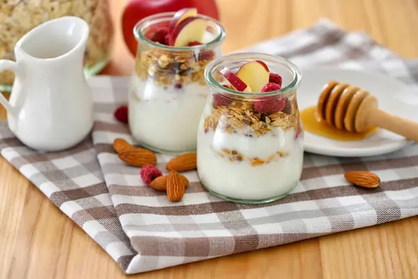 木のテーブルのガラス瓶にヨーグルト リンゴ ナッツ 蜂蜜が付いているクランチ グラノーラ 健康的な朝食のコンセプト ストック画像