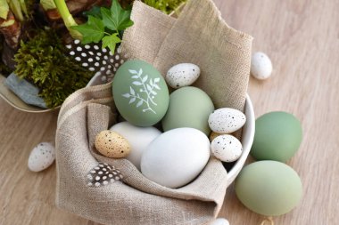 Süslü paskalya yumurtaları beyaz bir kâse içinde açık ahşap bir masanın üzerinde keten kumaş üzerinde. Üst görünüm