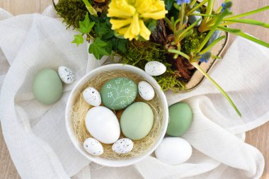 Süslü paskalya yumurtaları beyaz bir kâse içinde ve bahar çiçekleri keten kumaşın üzerinde. Üst Manzara. Mutlu Paskalyalar