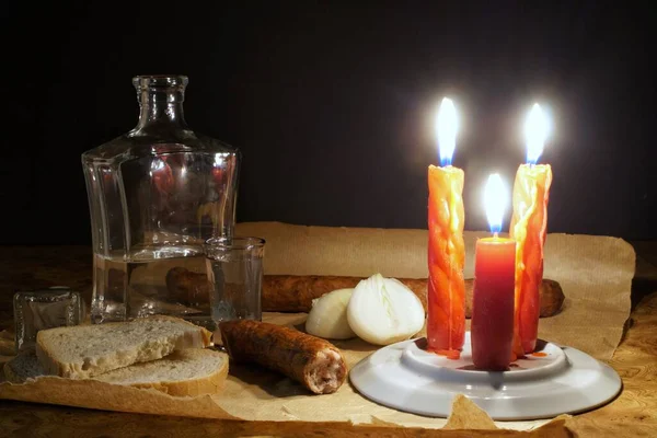 Gri kağıtta duran sosis, ekmek ve soğan. Yanında bir sürahi ve bir bardak dolusu votka var. Sahne üç mum ışığıyla aydınlatılır..