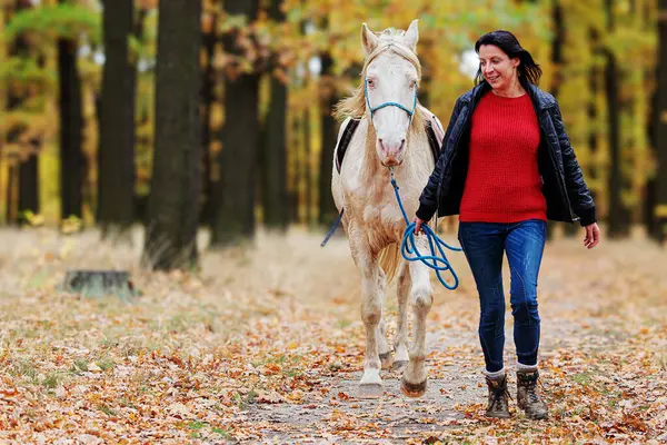 Young woman and a white horse walking together along the forest path