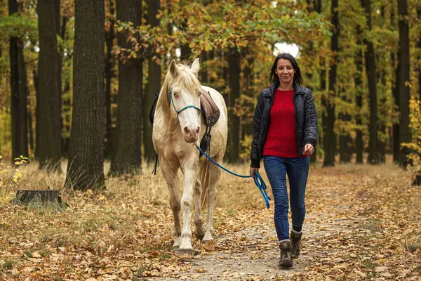 Young woman and a white horse walking through the autumn colored forest