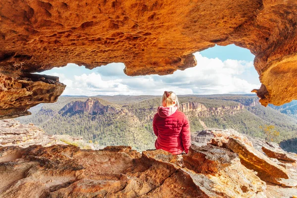 女性は壮大な山の景色を望む砂岩の洞窟に座っています ロイヤリティフリーのストック画像