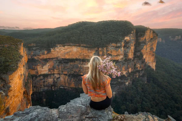 女性は岩の多い山の崖の上の端に座ってリラックスして美しい空の夕日を見ています 風に吹かれてネイティブの花を持っています ストック写真