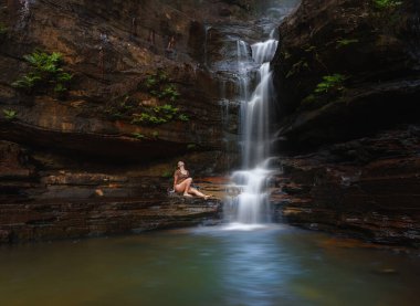 Dağın kenarındaki şelalede ve sığ havuzda oturan bir kadın.