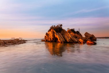 Rocky yıpranmış kumtaşı adası, gökyüzünde hafif bulutlarla altın güneş ışığıyla aydınlandı.