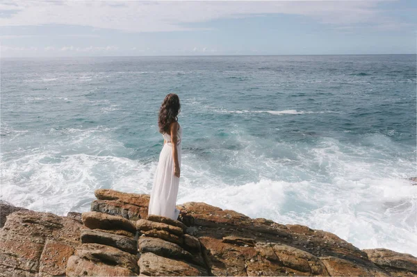 崖の上に立つ素晴らしい白いドレスで美しい花嫁の空中ドローンビューと海を見て 嵐の波と豪華な女性モデルと叙事詩シーン — ストック写真