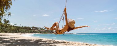 Siyah mayo ve hasır şapkalı Bikini plaj kızı yaz tatillerinde tropikal deniz kıyısında palmiye ağacında salıncakta sallanırken eğleniyor. Metin için kopyalama alanı olan yüksek kaliteli afiş fotoğrafı. 