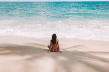 Kırmızı mayo giymiş, tropikal deniz kıyısında palmiye ağacının gölgesinde dinlenen, beyaz kumlu, okyanusa bakan bikinili kadın. Yaz tatili boyunca mükemmel bir modelin dikiz aynası. Yüksek kalite