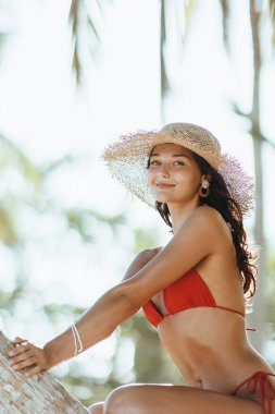 Summer lifestyle funny portrait of cute young bikini girl with tanned perfect body in red swimsuit, posing sitting on palm tree and looking at the camera at the tropical beach resort during sea